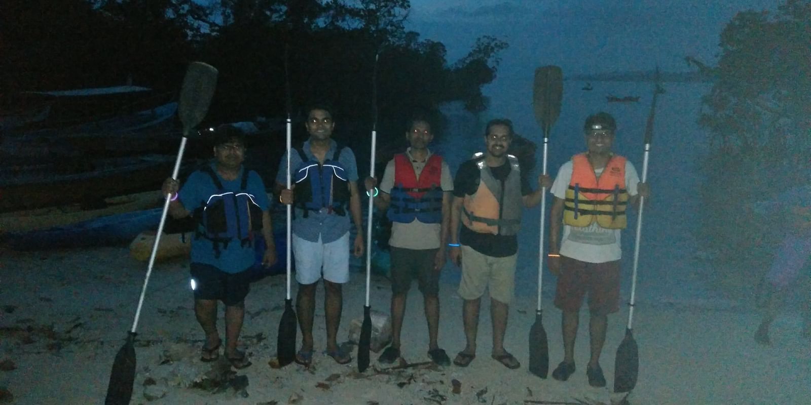 Five friends stand on a beach at dusk, holding paddles, wearing life jackets, and ready for an early morning kayaking trip.