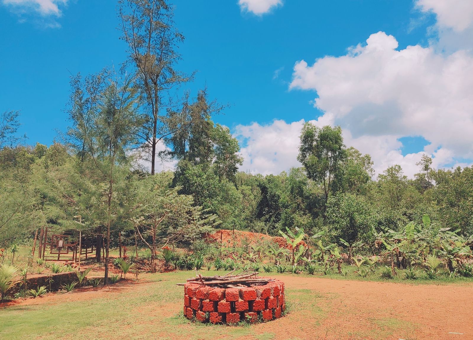Open garden with red-painted circular well arranged in a trees, and blue sky with clouds at Paradise Wild Hills Resort, Kollur.