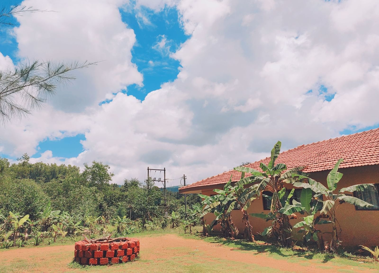 A wide outdoor view of a tiled-roof house surrounded by greenery with a circular arrangement of red-painted well in the garden at Paradise Wild Hills Resort, Kollur.