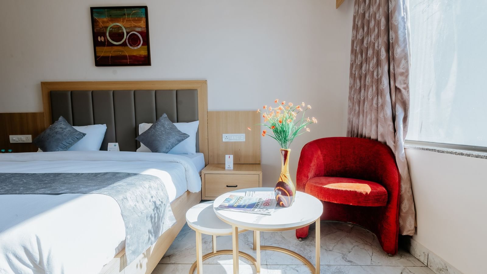 A brightly lit corner of a bedroom at Daksh The Grand Gowardhan, Dwarka, featuring a bed, a red armchair, and a small coffee table.