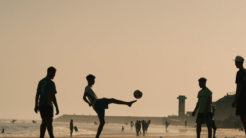 Silhouettes of a group of people, a football kicked high in the air, and a flat sandy beach are captured during a late afternoon.