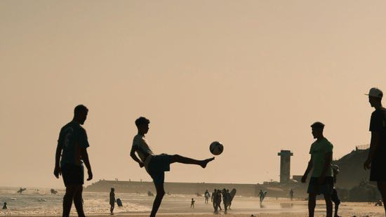 Silhouettes of a group of people, a football kicked high in the air, and a flat sandy beach are captured during a late afternoon.