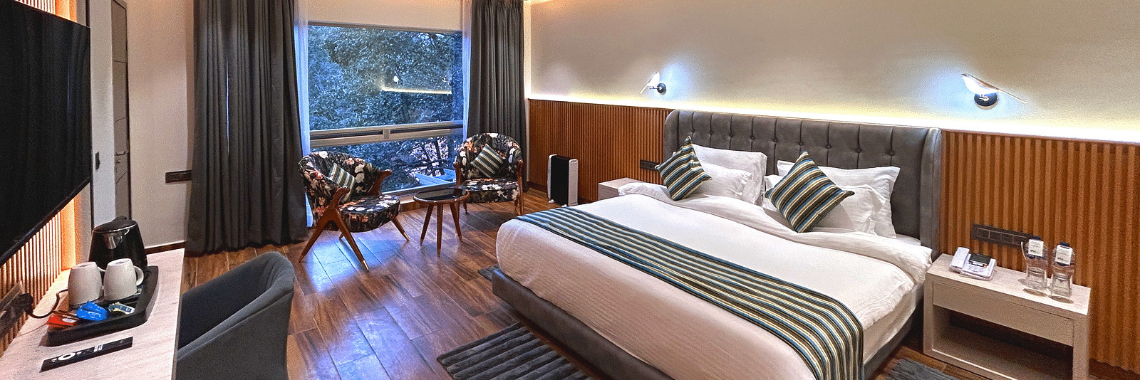 The premium room of DLS The Emerald, Nainital with a bed, bed side table, chairs, coffee table, a window overlooking the view, with warm lighting