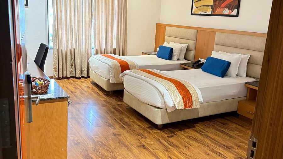 A corner view into a Deluxe Twin room at Kyriad Hotel, Gulbarga, showcasing two made beds with colorful accents, hardwood flooring, and a partial view of the room's artwork.