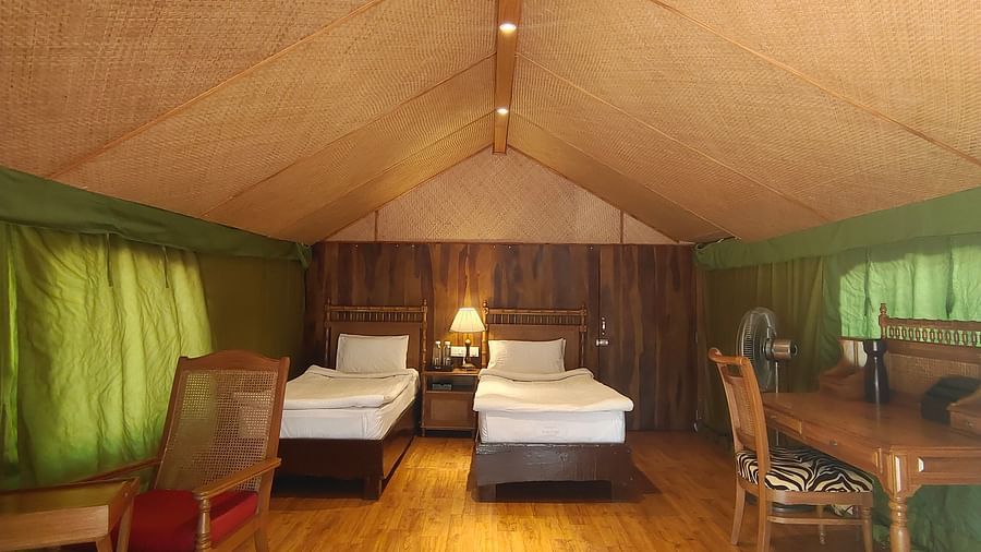 A zoomed out image featuring a cozy room with two beds inside and wooden flooring, lights lit and a lamp next to the beds.