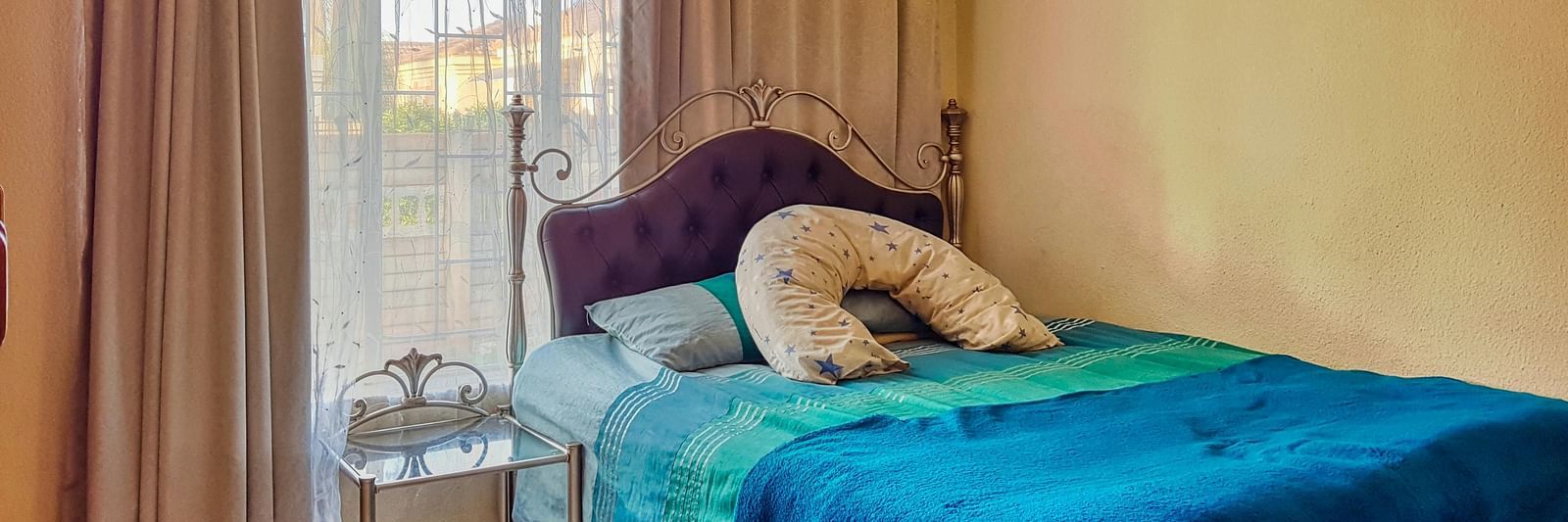 A bedroom featuring a double bed with blue sheets spread neatly over it with beige curtains covering the window behind it.