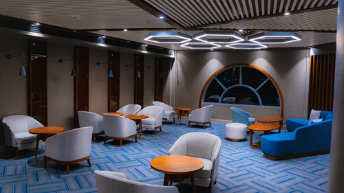 A wide angle shot of our restaurant in Hargeisa featuring round wooden tables, cushioned chairs, and bright ceiling fixtures - Serene Sarovar Premiere Hargeisa