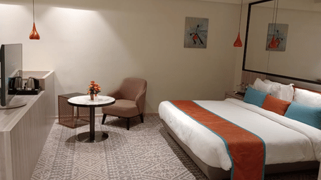 a contemporary king size bedroom with a lounge area, a TV, bedside table, and a mirror just above the bed frame at Lords Inn Rajkot