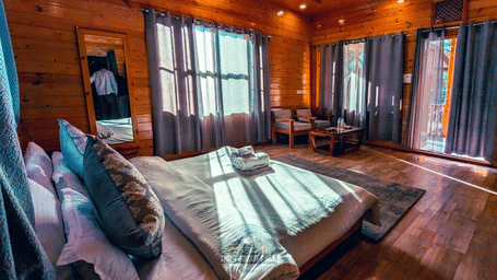 A Deluxe Room at Dawat e Drung by FrozenFall Resort, Drung, Tangmarg, with a double bed, wooden walls, window curtains, and a small seating area.