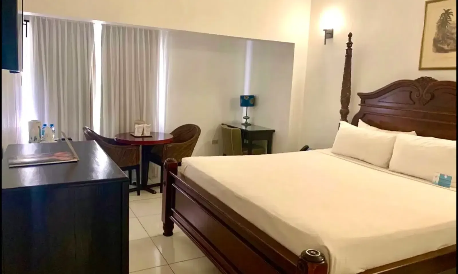 A well-lit hotel room features a large wooden bed with white linens, a dark dresser, and a seating area near a window | Executive King Room | Altamont Court, Jamaica