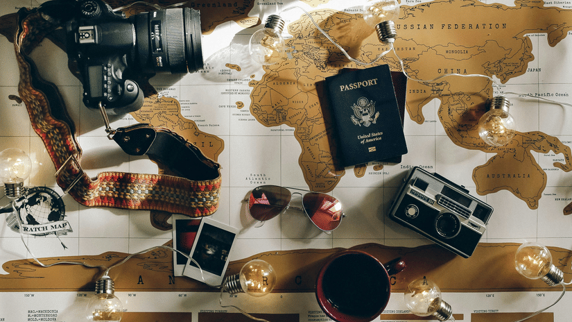 A flat-lay arrangement of travel items including a DSLR camera, a vintage camera, a passport, and photos on a world map.