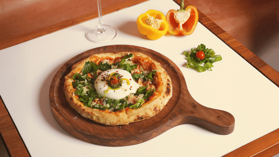 A freshly baked pizza served on a wooden board at The Bakehouse topped with burrata cheese fresh herbs and greens alongside a glass of red wine and bell peppers on a white table.