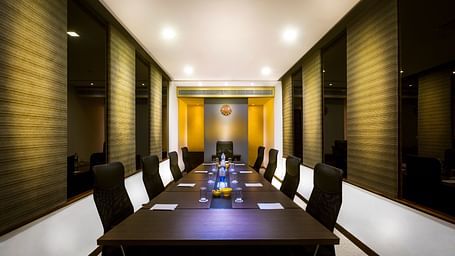 Modern meeting room with a long, rectangular table surrounded by black leather chairs. The room has neutral walls and ample lighting.