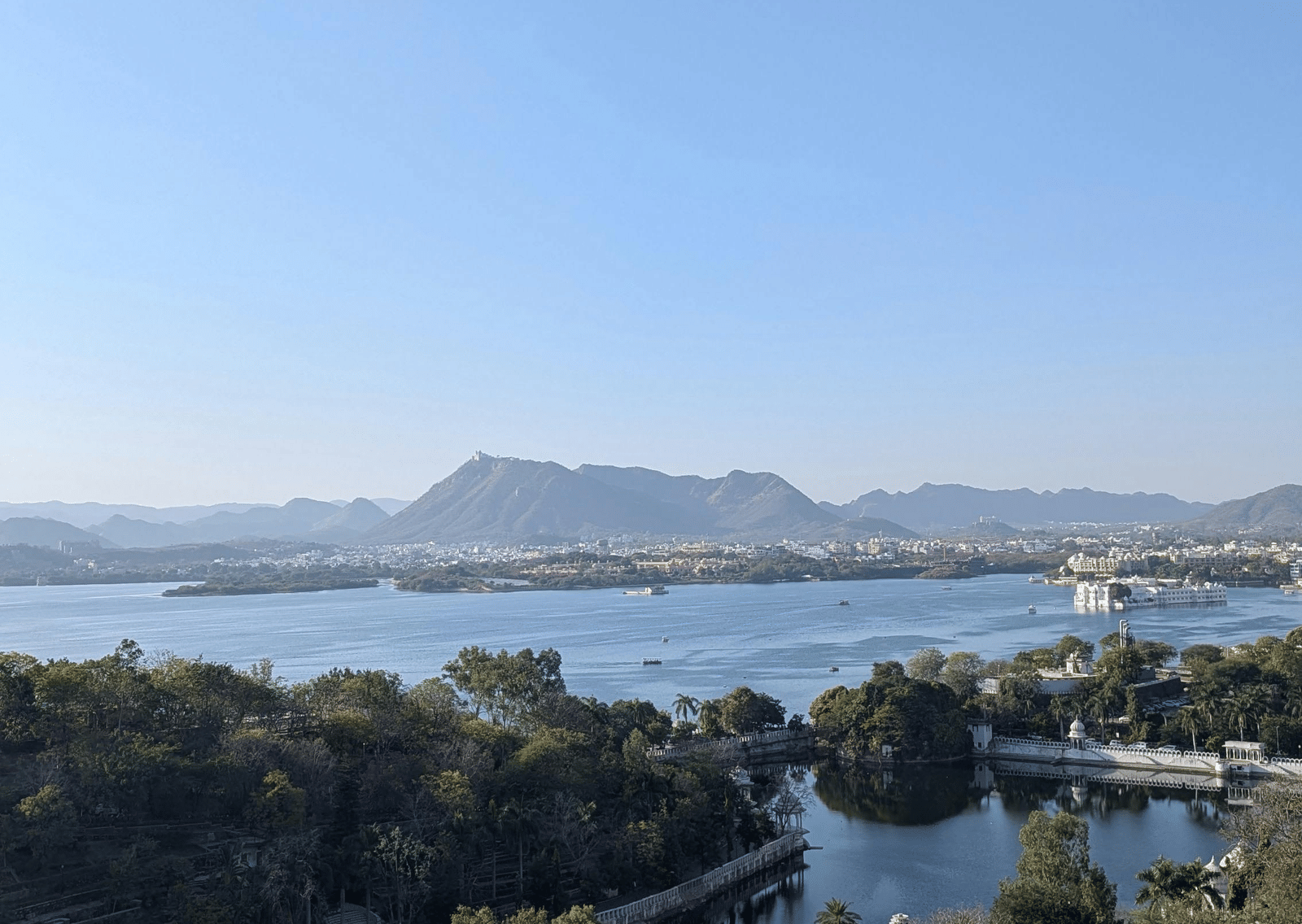 A wide-angle landscape view of a large, serene lake surrounded by green trees and scrub, with distant hills under a bright blue sky.