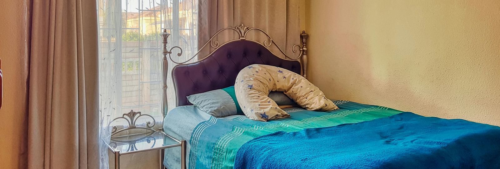 A bedroom featuring a double bed with blue sheets spread neatly over it with beige curtains covering the window behind it.