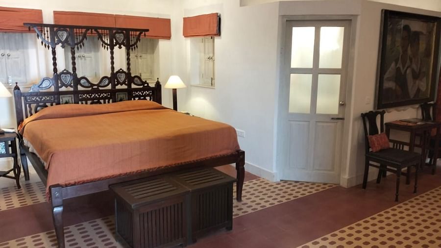 A bedroom with a four-poster bed with an orange bedspread, a wooden chest at the foot of the bed, a doorway, and a seating area with chairs and a table with artwork on the wall at Tijara Fort-Palace - 19th Century, Alwar.