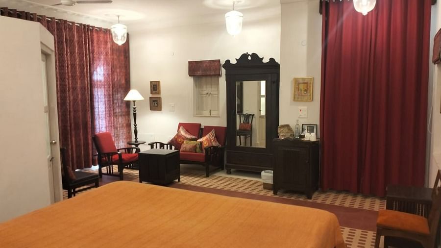 A bedroom featuring a bed with an orange bedspread, a dark wooden wardrobe with a mirror, seating area with chairs and a table, and curtains on the windows at Tijara Fort-Palace - 19th Century, Alwar.