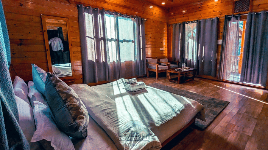 A Deluxe Room at Dawat e Drung by FrozenFall Resort, Drung, Tangmarg, with a double bed, wooden walls, window curtains, and a small seating area.