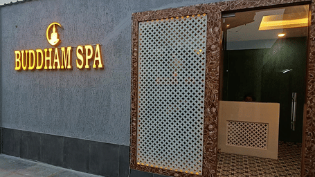Exterior entrance of Buddham Spa, featuring a grey textured wall with a golden sign and an ornate wooden-framed window lattice screen.