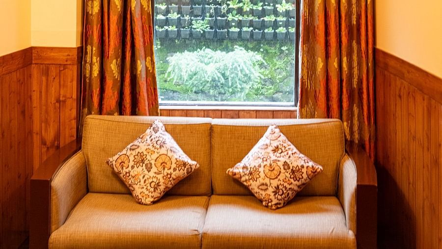 A sitting area in the Oak Room features a beige sofa with patterned cushions, a large window with ornate drapes, and a wooden ceiling at The Pavilion, Nainital