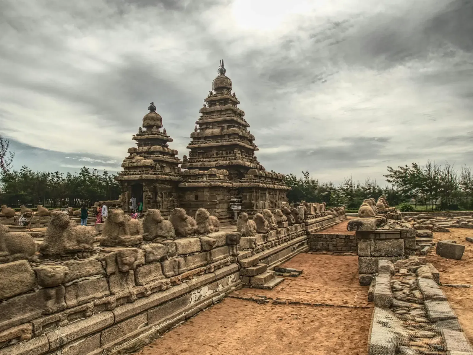 A wide-angle view of the Shore Temple complex, showing the ancient stone structures and a pathway leading towards them under a cloudy sky.