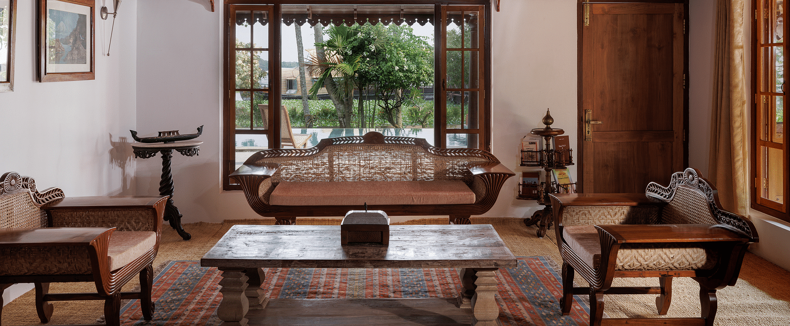 A shot of a room showcasing the sitting area with wooden couches, a coffee table, and a huge window overlooking the view - BluSalzz Villas - Muhamma Resort, Alleppey
