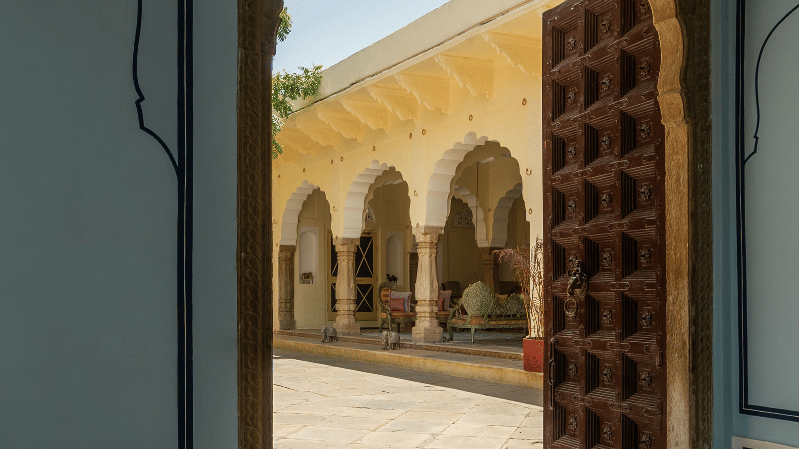 A carved wooden door stands slightly ajar, offering a view into a courtyard with arched passages at Fort Barli, Ajmer, framed by decorated interior walls.