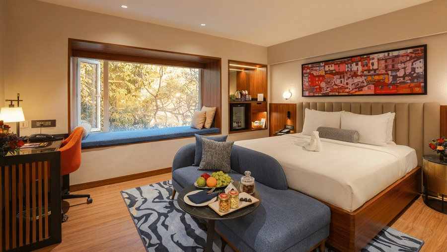 Pride Premier Pune hotel room featuring a double bed, blue sofa, window seat, work desk, and a colourful painting on the wall.