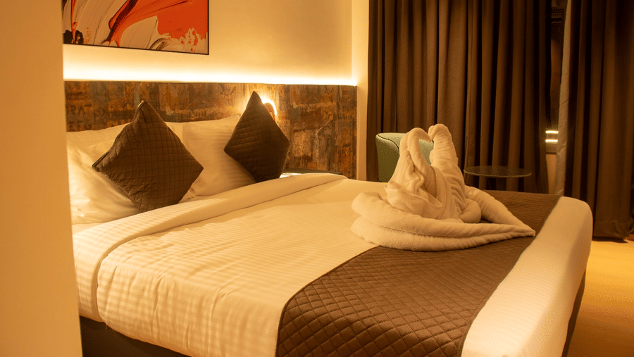 A hotel bedroom with king size bed, pillows, bedside lighting, and folded towel decor on the bed.