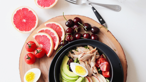 A bowl of salad surrounded by grapefruit, cherries, and tomatoes with a fork and a spoon next to it