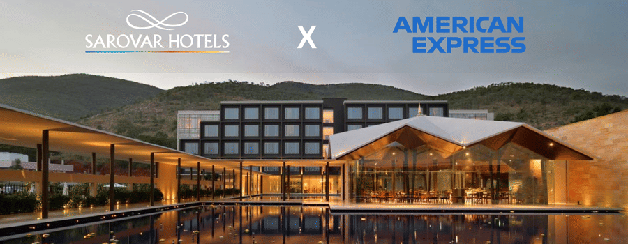 A building with several windows, restaurant with glass walls, and a corridor with plants and lights. A promotion for Amex Card holders at Sarovar Hotels.