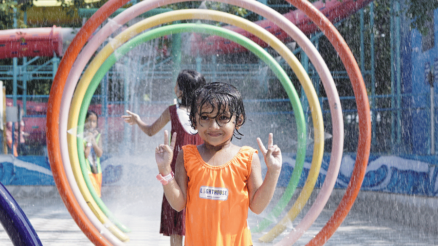 A young girl is standing beneath a rainbow-like water spray structure in a shallow pool area at Lighthouse Waterpark & Resort, Nagpur.
