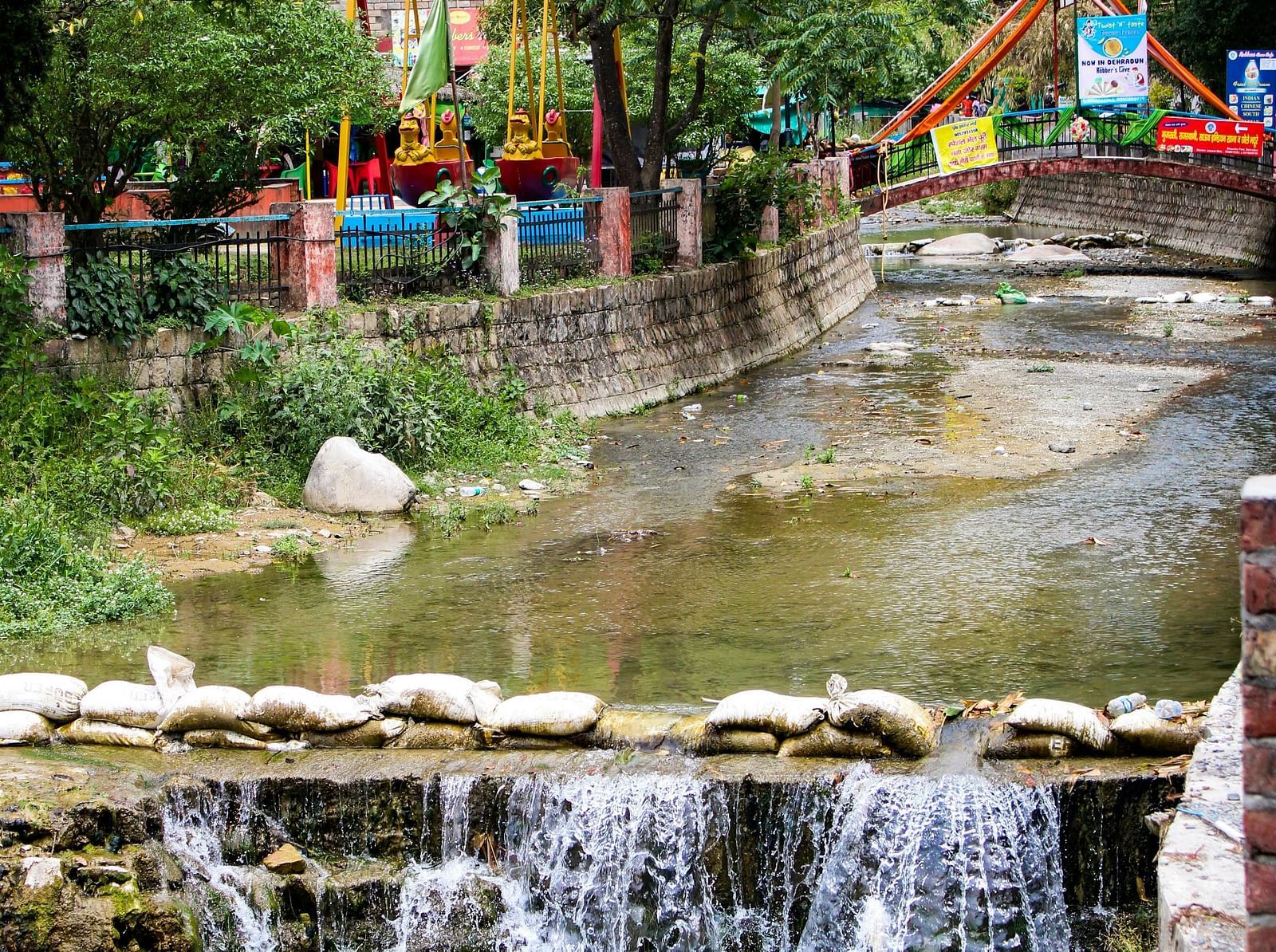 A small, playful waterfall flowing under a pedestrian bridge in a park, with surrounding greenery and recreational structures.