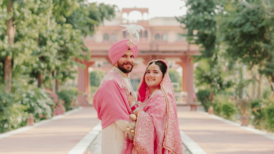 A vertical portrait of a bride and groom dressed in coordinated pink and cream wedding outfits standing on an outdoor pathway at Umaid Palace.