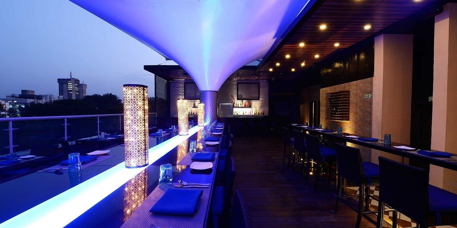 Skylit Rooftop Lounge and Bar at The Cunningham Hotel featuring ambient blue lighting, elegant seating, a modern bar, and a city skyline view at dusk