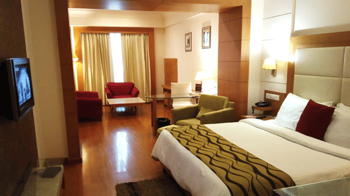 A Junior Suite at Ambrosia Sarovar Portico, Haridwar with a bed, seating area, a wall-mounted TV, wooden flooring, and curtains separating the living area.