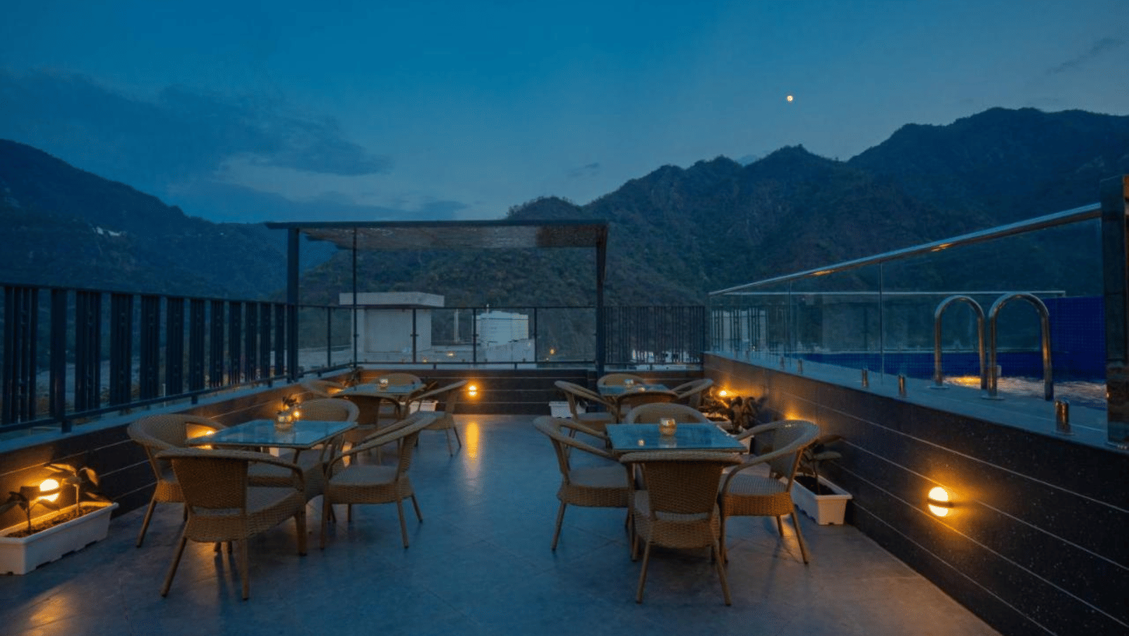 A dimly lit outdoor rooftop restaurant area with several tables and chairs, and a pergola, set against a backdrop of mountains under a twilight sky at Perfectstayz Premium Laxmi Heritage.
