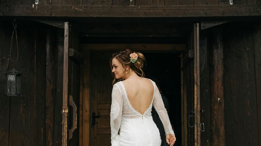 A bride in a white gown stands in a doorway of a rustic, dark-colored building, looking away