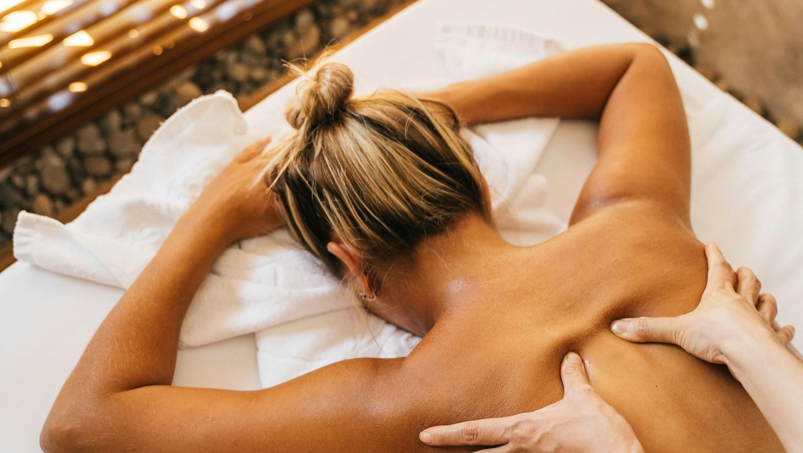 A top-down shot of a person receiving a back massage on a massage table, with the masseuse's hands applying pressure to their back.