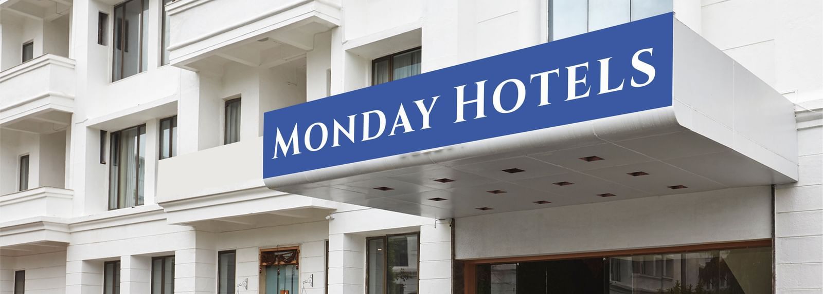 The exterior of a hotel building featuring a huge board on the top of the entrance reading 'Monday Hotels' along with a drive way in front of the building.