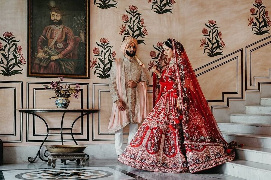 A bride and groom stand together indoors, with a portrait on the wall and floral designs behind them at  Umaid Palace - Best Luxury Heritage Resort Near Jaipur.  