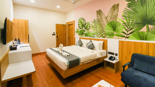 Modern hotel room with a double bed, tropical-themed wall art, wooden furnishings, a ceiling fan, and a comfortable seating area - MGM Vailankanni Residency.