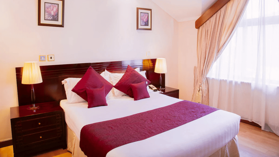 Standard Queen Room featuring a plush bed placed next to curtained window and wall hangings with lamps on both sides of the bed on nightstands at Boma Inn Nairobi