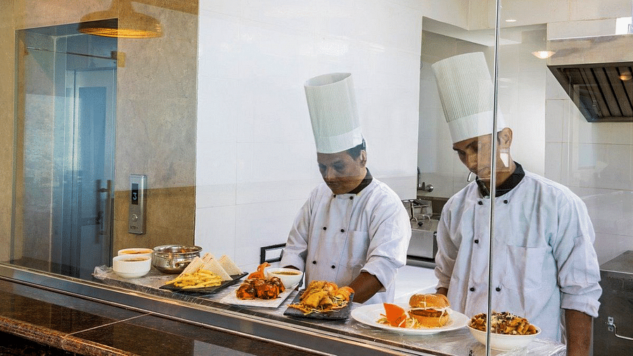 Two chefs preparing dishes in the kitchen as seen from the transparent glass partition at the restaurant at Regenta Inn DLS.