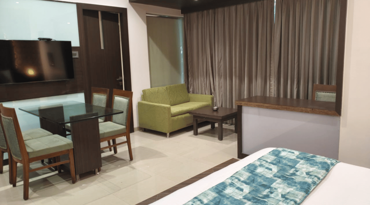 A bedroom layout at Hotel Mumbai House, Thane featuring a corner of a bed with a blue patterned runner, a glass top dining table with 2 upholstered chairs, a green fabric sofa, and a wall television.