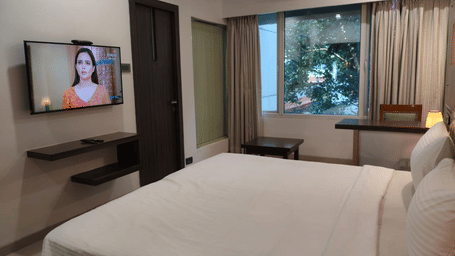 Wide view of a room at Hotel Mumbai House with a wall-mounted TV, single bed, large window, and lush greenery beyond.
