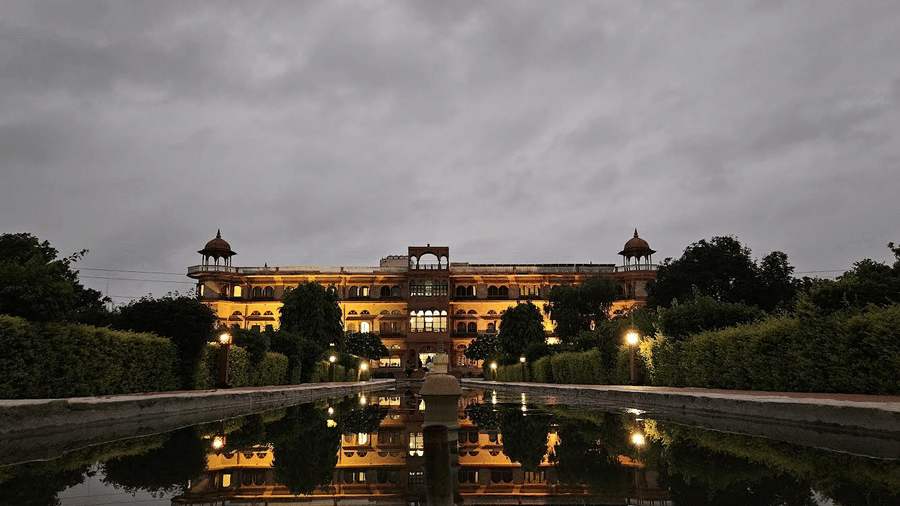 A well lit facade of Umaid Palace, a premiere resort near Bhangarh Fort, featuring stretched and multi storey building with lights on. The property is covered by trees and there is also a huge fountain in the front yard.