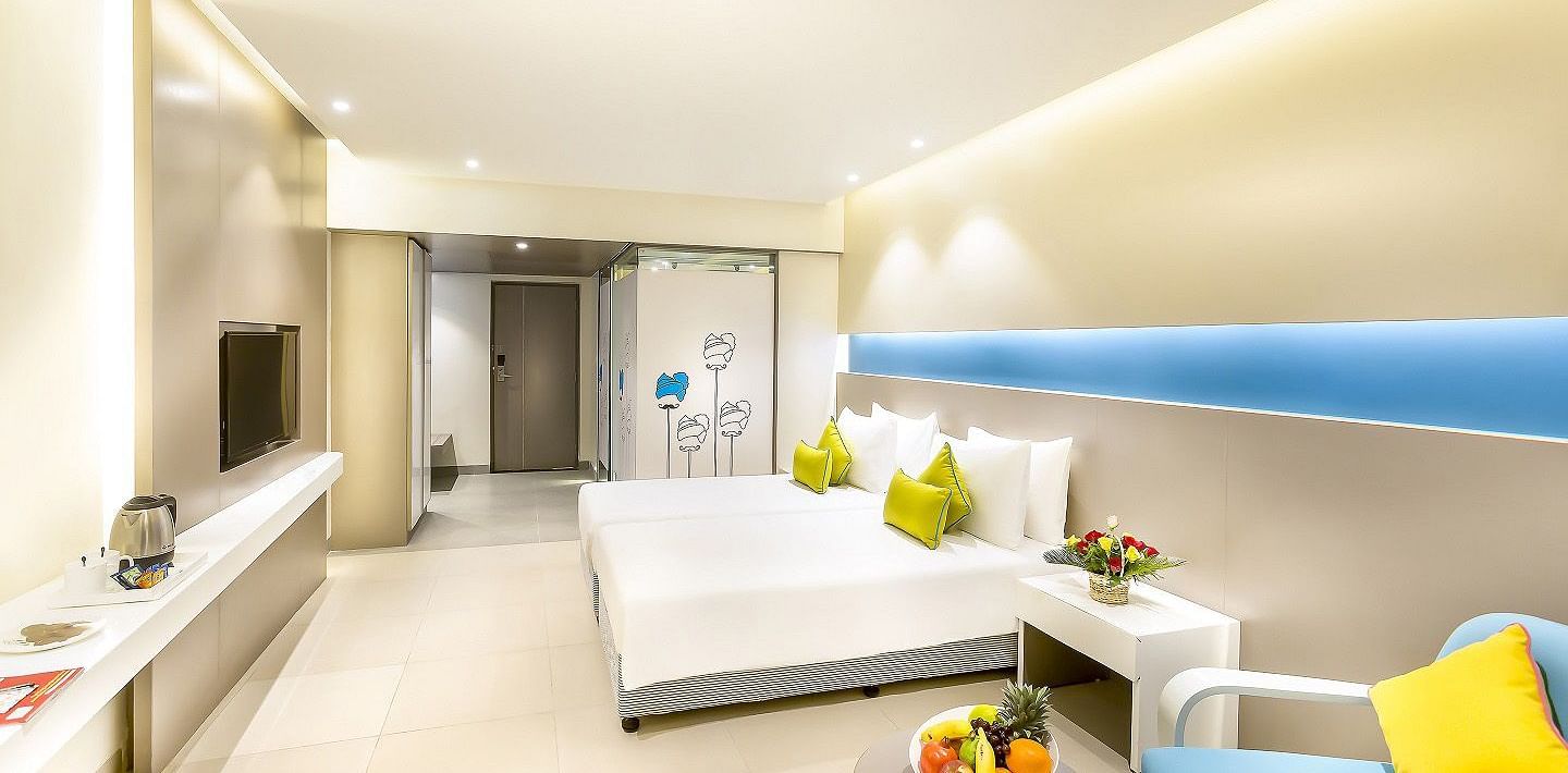 A spacious room features a large bed with a headboard stretching across the wall. Cushion chairs sit beside a wooden bedside table, and a wall-mounted television is positioned opposite the bed