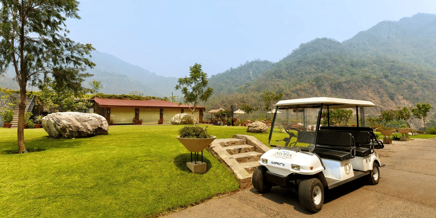 Scenic outdoor view at Aloha on the Ganges Resort with white golf cart on paved path, grassy area, decorative rocks, red-roof building, and misty green hills in the background.