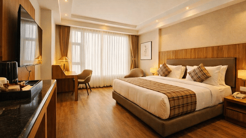 A bedroom at Pride Premier Darjeeling featuring a double size bed, a flat TV screen on the wall, a workstation with chair and desk, a sofa chair, and a large window with curtains drawn.
